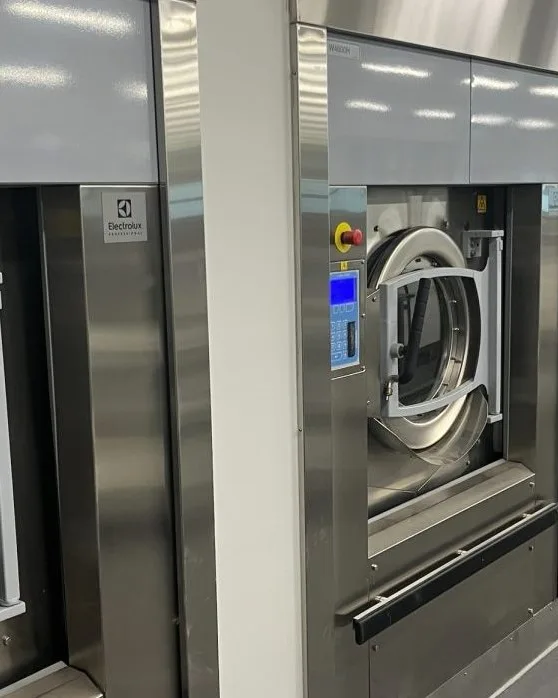 A large-capacity Electrolux on-premises washer with an open door, installed along a wall of similar machines