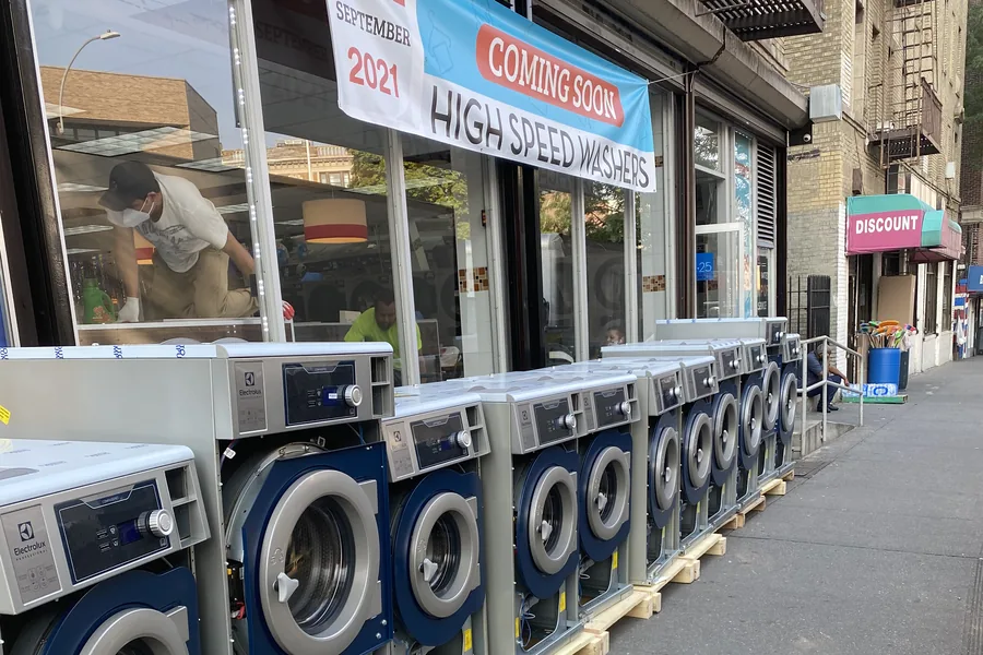 Row of new Electrolux washers on pallets on a New York City sidewalk, under a Coming Soon High Speed Washers banner